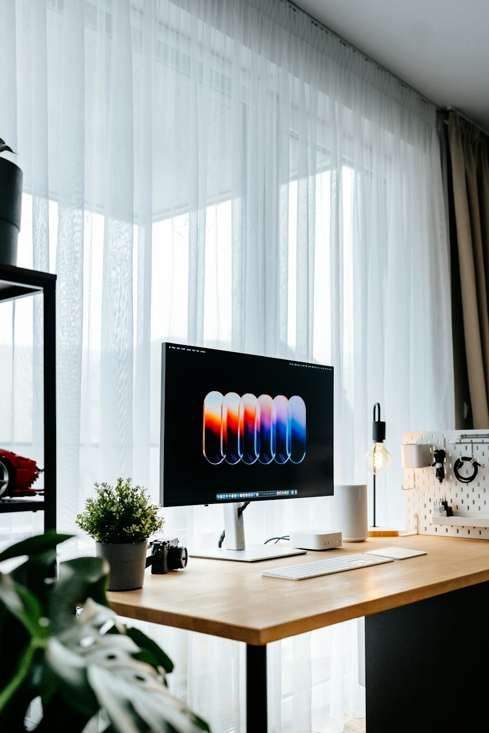 Monitor komputer dan keyboard di atas meja kayu terang dengan tanaman.