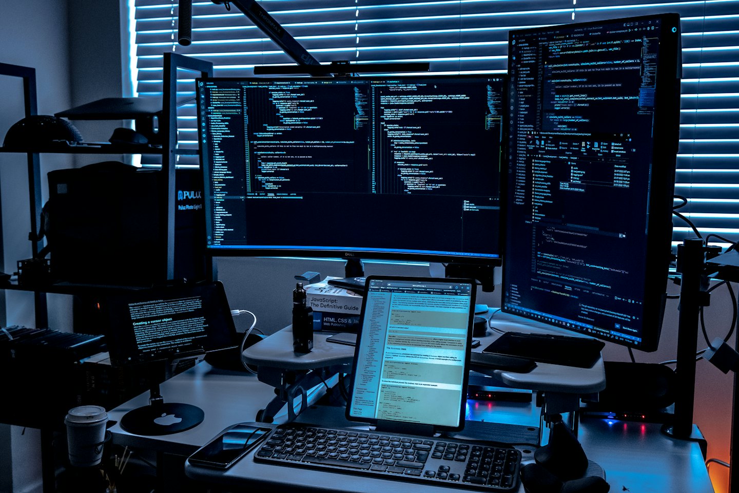 Dua monitor besar dan laptop menampilkan kode di ruang kerja yang gelap.